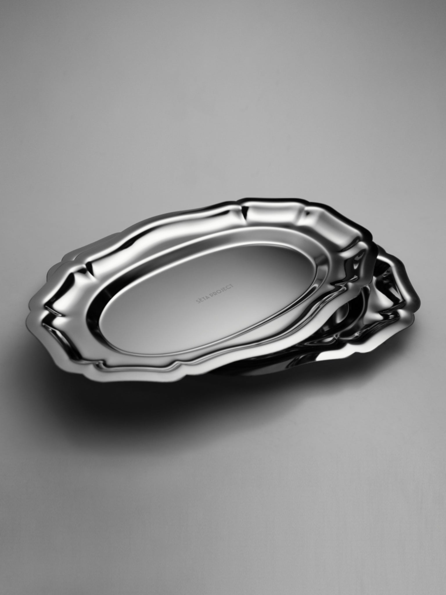Set of two polished stainless steel oval trays with scalloped edges and smooth surfaces.