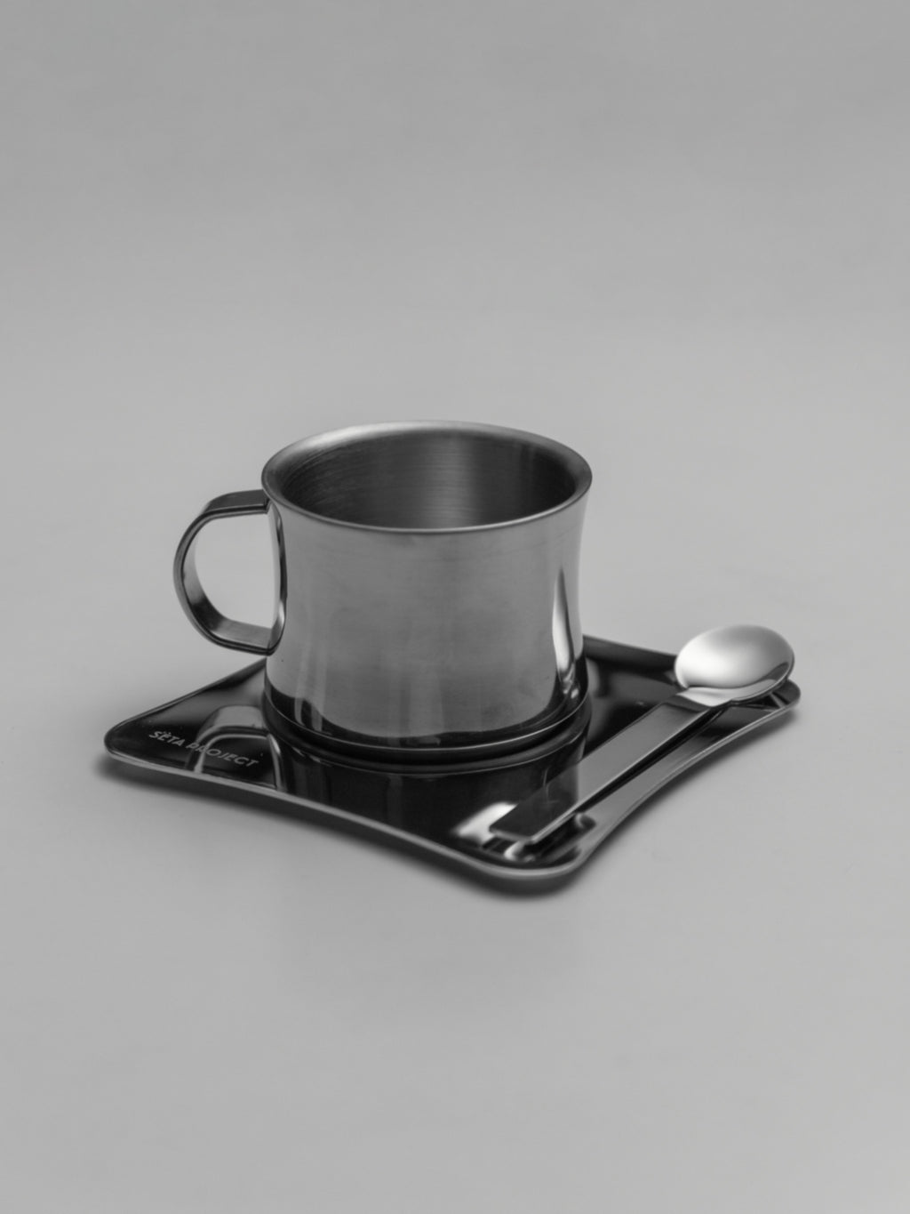 Stainless steel coffee cup with handle on black square tray with a small spoon.