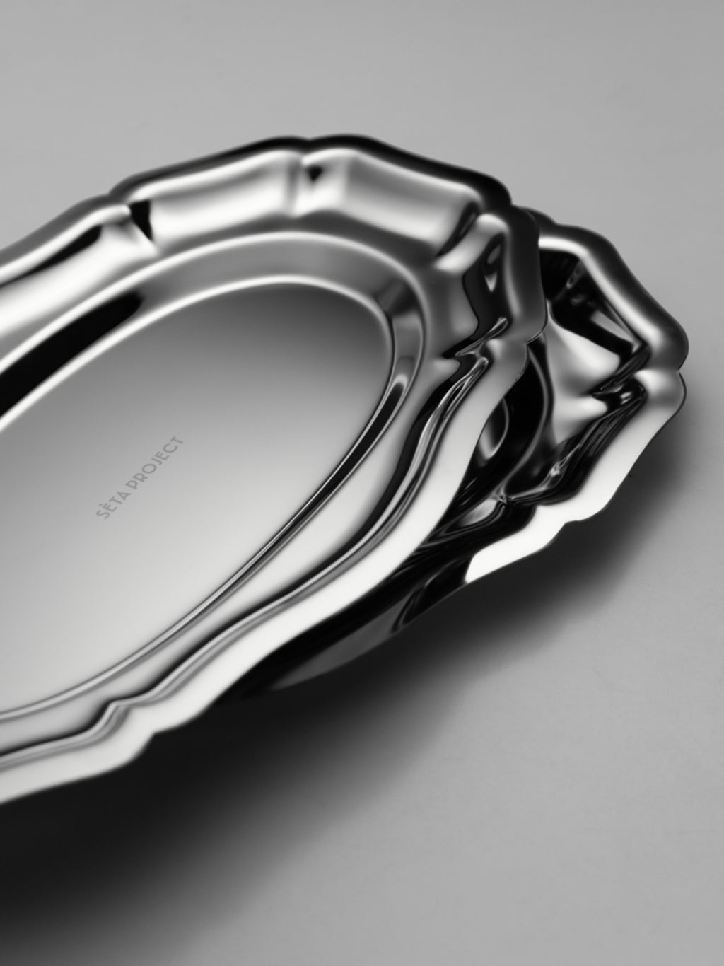 Stacked stainless steel CELENE trays with scalloped edges and a polished finish on a gray surface.