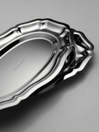 Stacked stainless steel CELENE trays with scalloped edges and a polished finish on a gray surface.