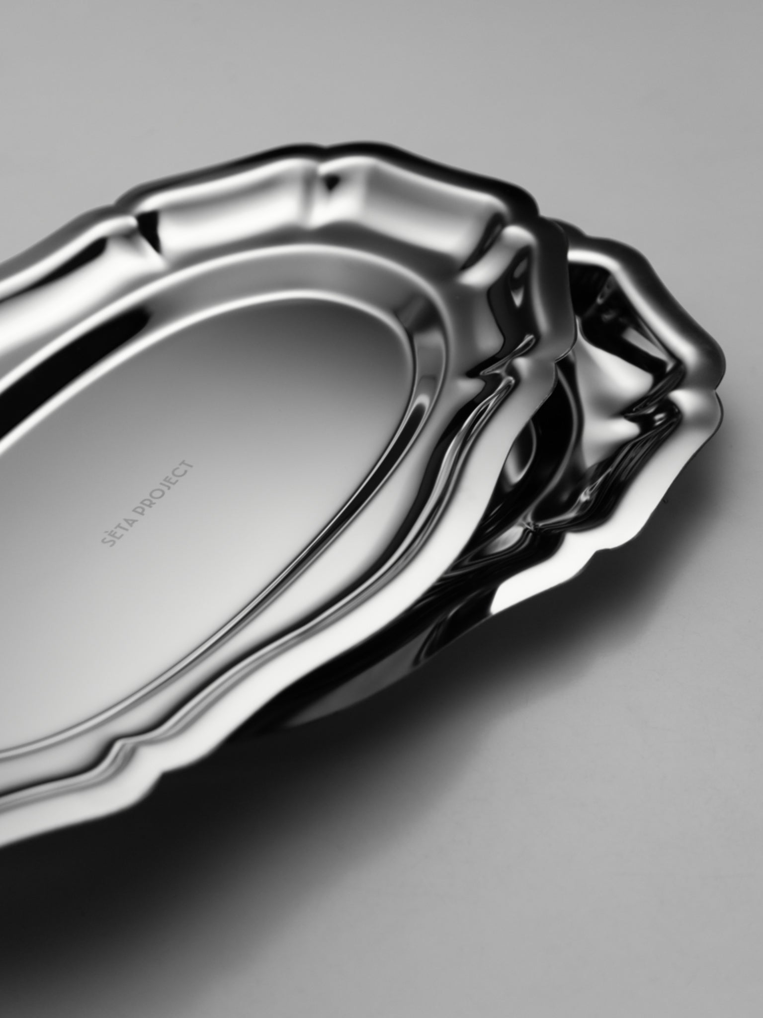 Stacked stainless steel CELENE trays with scalloped edges and a polished finish on a gray surface.