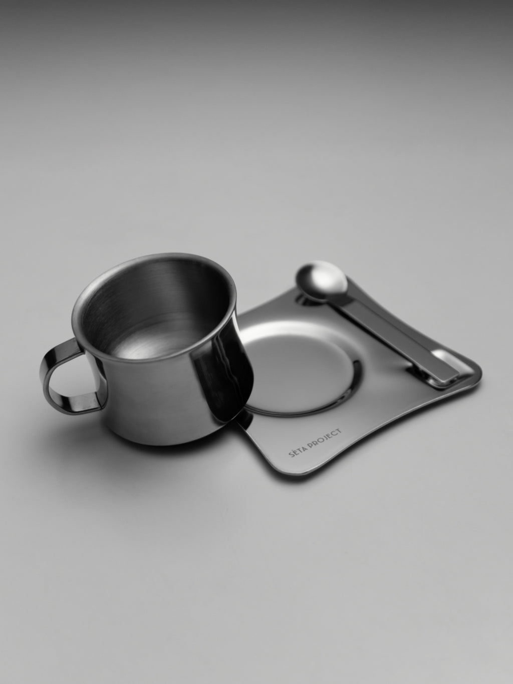 Stainless steel coffee cup with matching saucer and teaspoon on a gray surface.