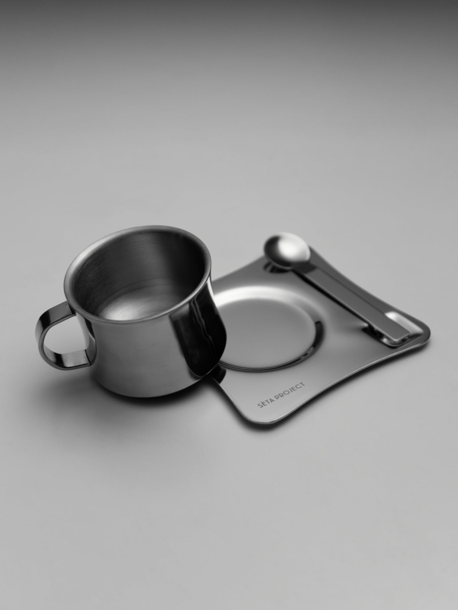 Stainless steel coffee cup with matching saucer and teaspoon on a gray surface.