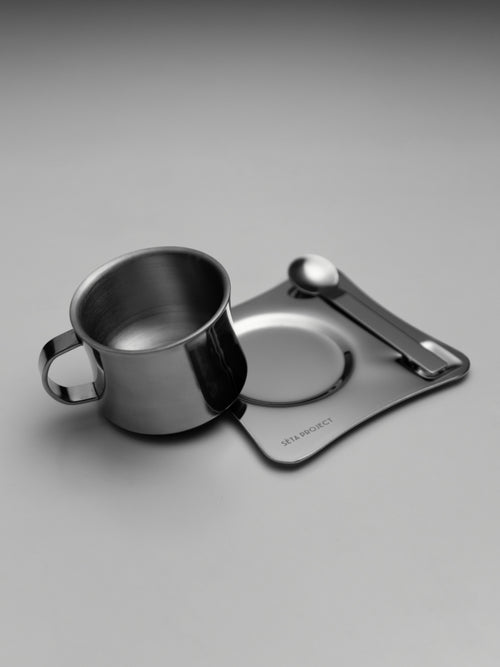 Stainless steel coffee cup with matching saucer and teaspoon on a gray surface.