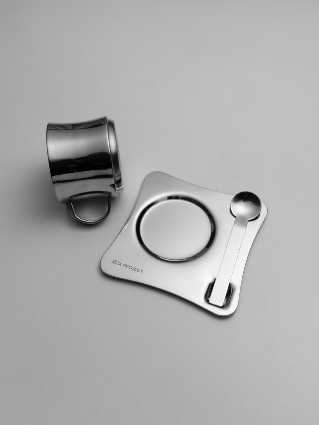 Polished stainless steel coffee cup with matching saucer and spoon on gray background.