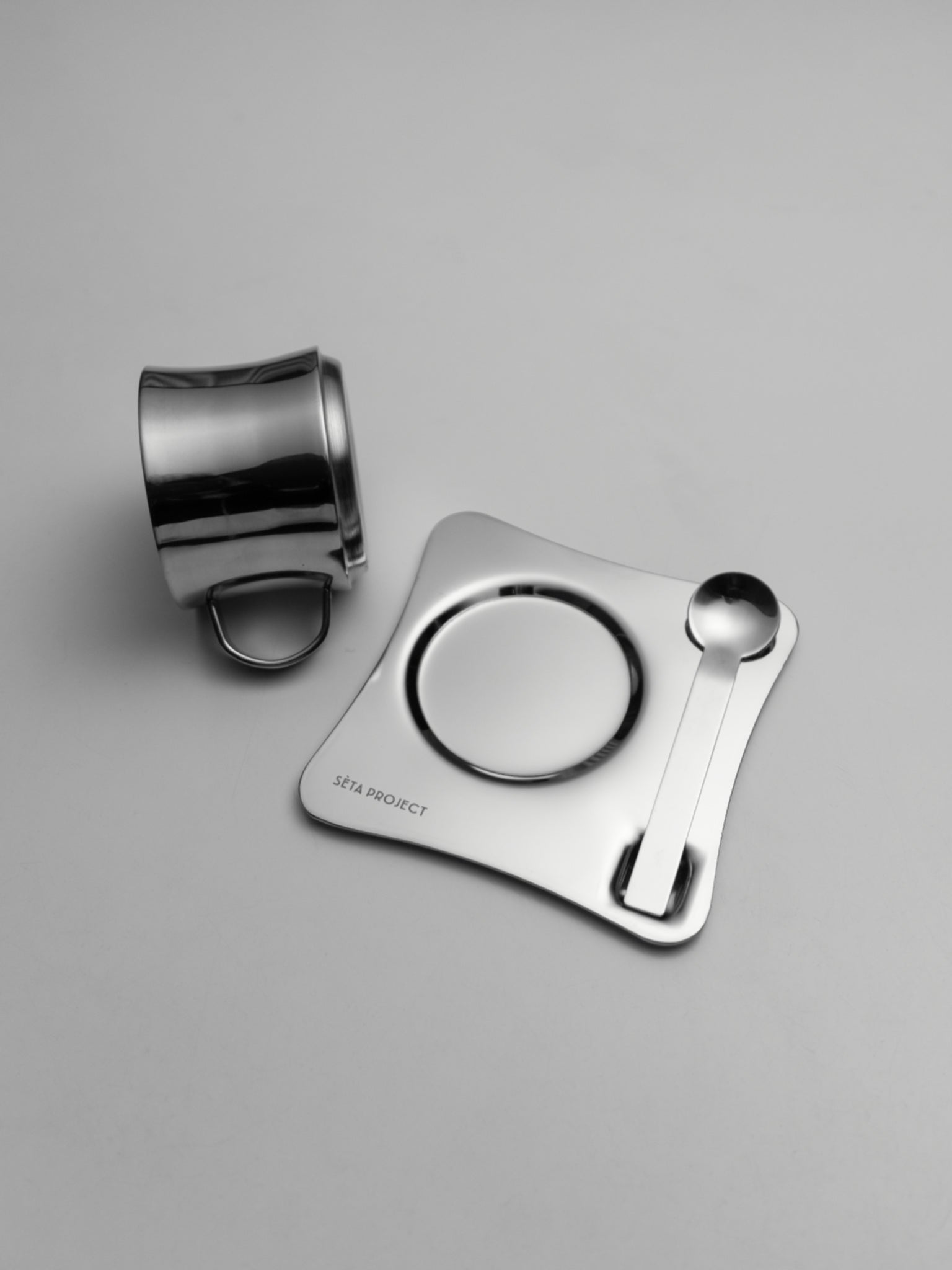 Polished stainless steel coffee cup with matching saucer and spoon on gray background.