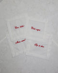 Set of four white linen cocktail napkin coasters each embroidered with red cursive phrases.