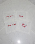 White linen cocktail napkin coasters with red embroidered phrases on a marble surface, set of four.