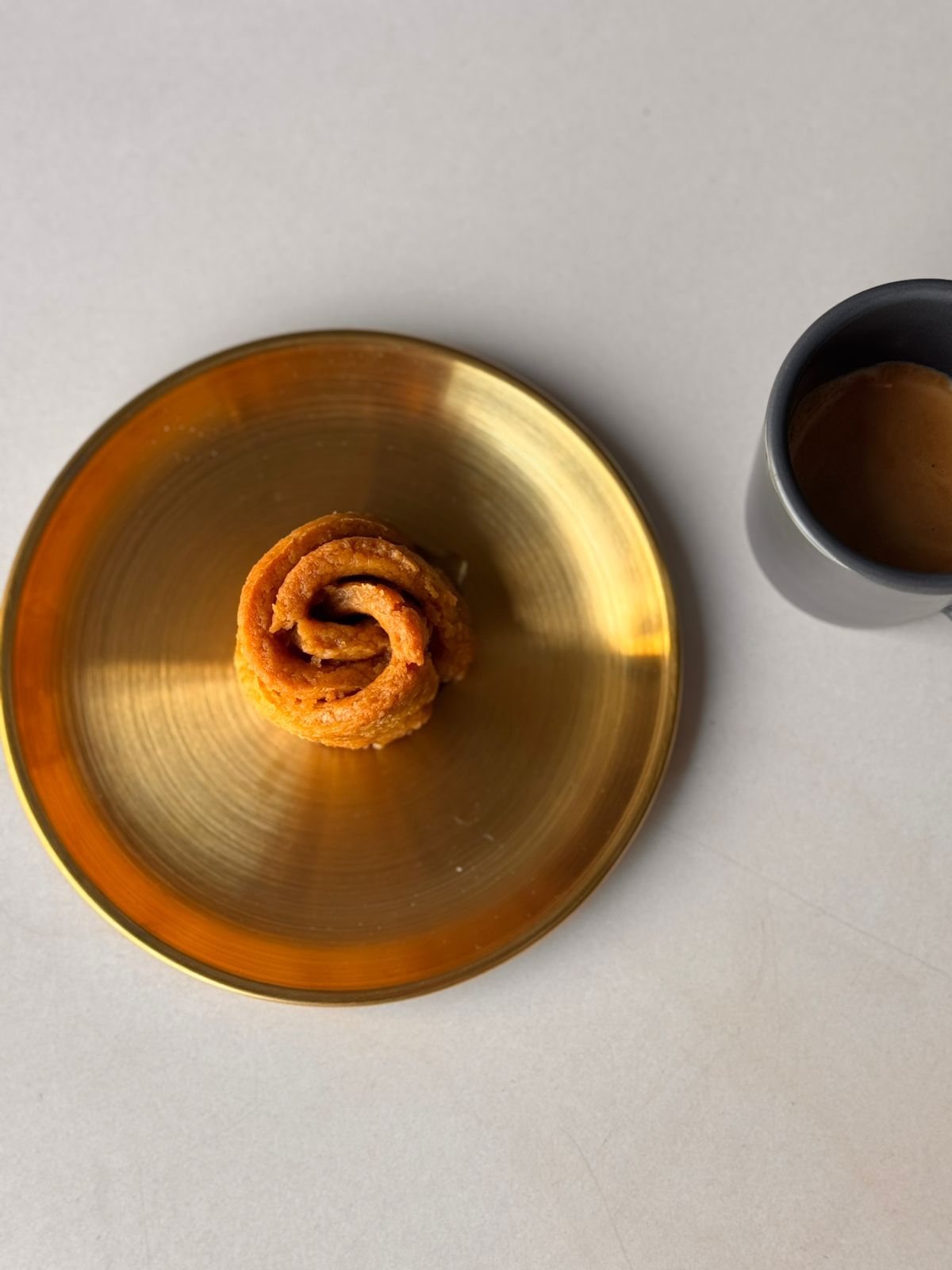 Small round gold plate with spiral pastry and black ceramic cup of coffee on light surface.