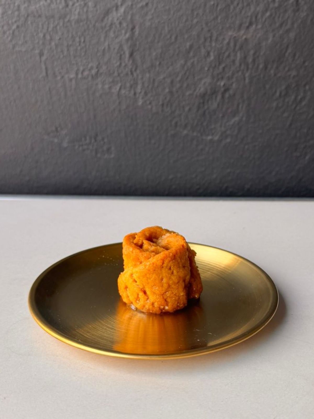 Petite 14cm round gold plate with a small orange dessert on top against a gray textured wall background.